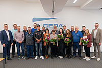 Eine Gruppe von 18 Menschen steht nebeneinander in einem Raum, einige halten Blumensträuße.