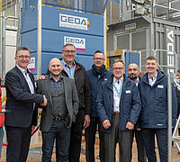Eine Gruppe von sieben Männern steht lächelnd zusammen vor einer Aufzugskonstruktion der Marke "GEDA".