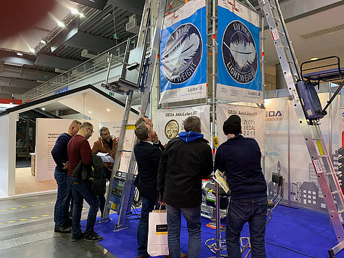 Personen stehen vor einem Messestand, betrachten aufmerksam eine Steigtechnik-Präsentation.