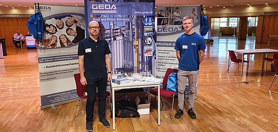 Zwei Personen stehen an einem Tisch mit Flyern vor einem Messestand der Firma GEDA, zu sehen sind Roll-Ups mit Texten über Aufzugstechnik.