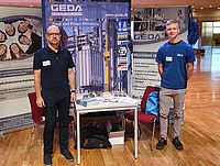 Ein Mann steht lächelnd an einem Messestand mit Broschüren und einem Roll-up von GEDA, das Aufzüge illustriert.
