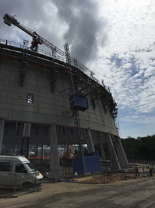 Gerüstetes Betongebäude im Bau mit Kran oben; Arbeiter und Lieferwagen im Vordergrund; wolkiger Himmel im Hintergrund.