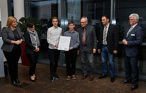 Eine Gruppe von sieben Personen steht nebeneinander; zwei Jugendliche halten ein Zertifikat.