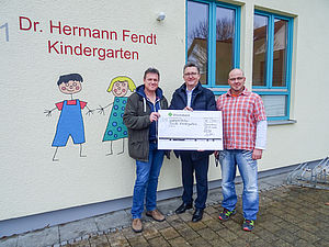 Drei Männer halten einen großen Scheck vor dem Dr. Hermmann Fendt Kindergarten, zwei Fenster im Hintergrund.