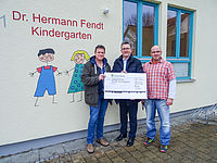 Drei Männer halten einen großen Scheck vor dem Dr. Hermmann Fendt Kindergarten, zwei Fenster im Hintergrund.