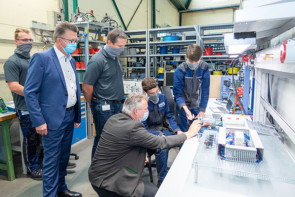 Mehrere Personen mit Masken prüfen Kabel und Technikgeräte auf einem langen Tisch in einer Werkstatt.