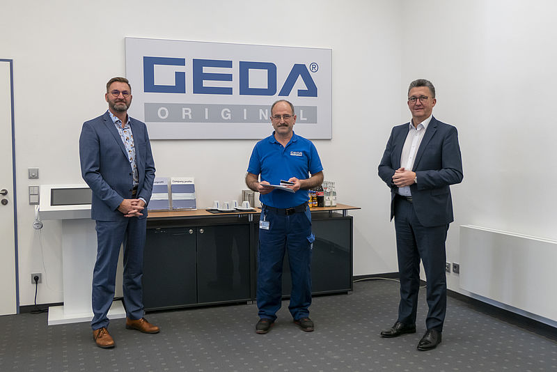Drei Männer stehen in einem Büro vor einem Schild mit der Aufschrift "GEDA ORIGINAL.