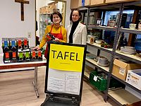 Zwei Frauen stehen hinter einem Schild mit der Aufschrift "Tafel" und neben einem Tisch mit Saftkartons, in einer Vorratskammer mit Regalen voller Lebensmittel und Geschirr.
