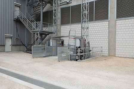 Aufzug steht still auf grauem Industriegelände neben einem Gebäude mit Metalltreppen.