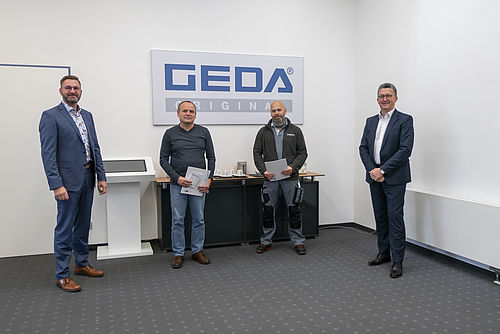 Vier Männer stehen in einem Büro vor einem Schild mit der Aufschrift "GEDA ORIGINAL".