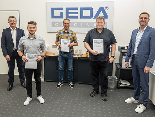 Fünf Männer stehen in einem Büro vor einem Schild mit der Aufschrift "GEDA ORIGINAL", einige halten Urkunden.