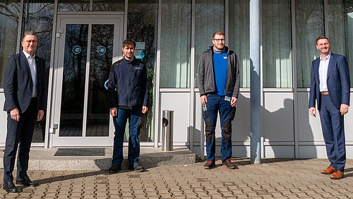 Vier Männer stehen vor einem modernen Gebäude mit Glastüren.