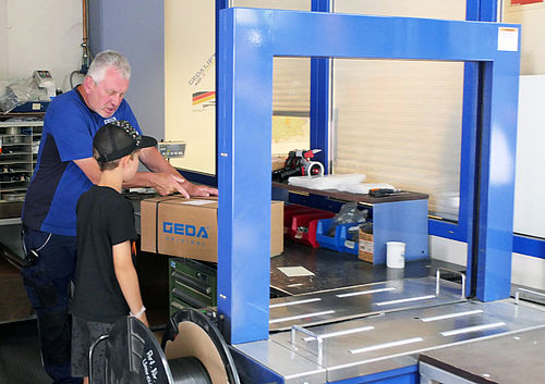 Ein Mann packt mit einem Jungen einen Karton der Marke "GEDA" an einer blauen Verpackungsmaschine.