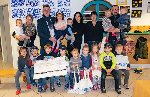 Erwachsene halten Kinder, während eine Gruppe von Kindern auf dem Boden sitzend einen großen Scheck und Geschenke präsentieren.
