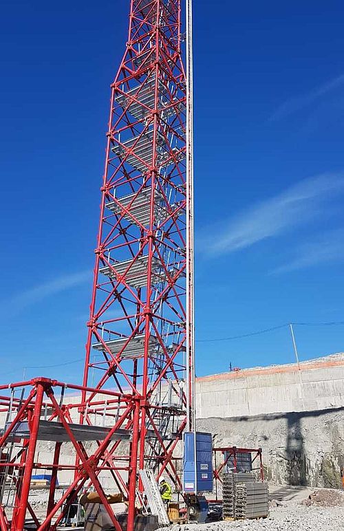 Ein roter Metallturm steht auf einer Baustelle, umgeben von Betonmauern und blauem Himmel.
