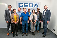 Acht Personen stehen nebeneinander in einem Büro, vor einem großen Schild mit der Aufschrift "GEDA".