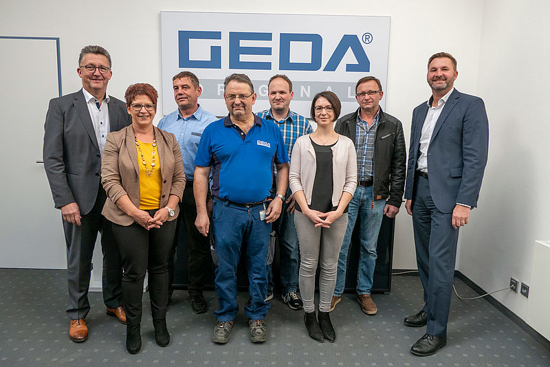 Acht Personen stehen nebeneinander in einem Büro, vor einem großen Schild mit der Aufschrift "GEDA".