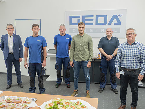 Sechs Männer stehen nebeneinander in einem Raum mit einem „GEDA Original“-Schild an der Wand; vor ihnen liegt ein Tisch mit belegten Brötchen.