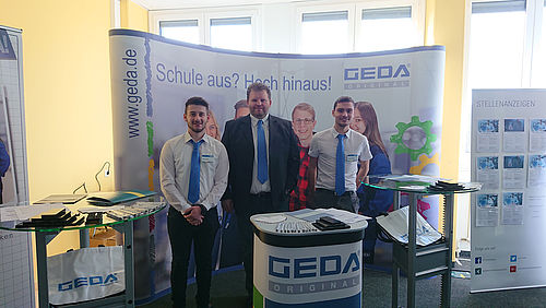 Drei Männer mit blauer Krawatte stehen lächelnd vor einem Messe-Stand.