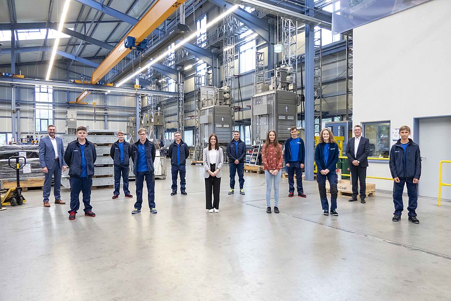 Mehrere Personen stehen in Arbeitskleidung und Business-Outfits nebeneinander in einer großen Industriehalle verteilt.